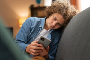 Žmogus, naudojantis išmanųjį telefoną, jaučiasi pavargęs nuo informacijos pertekliaus, simbolizuojantis technologijų poveikį psichinei sveikatai.