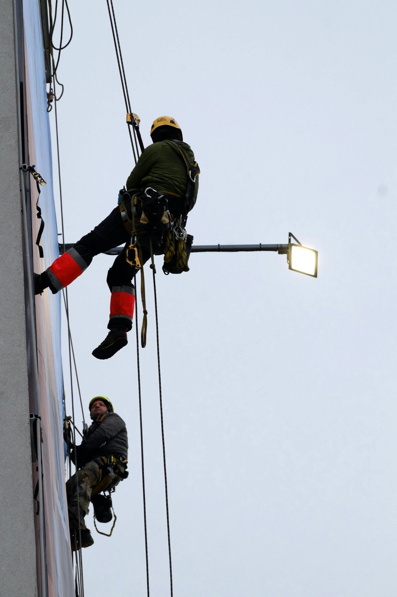 Darbuotojas su pilna saugos ekipuote ir šalmu dirba aukštyje ant metalinių konstrukcijų.